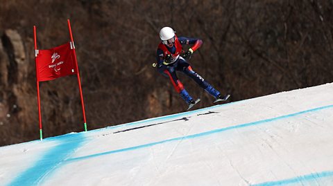 Skier Neil Simpson goes past a red banner reading Beijing 2022. Both skies are off the ground as he flies around the corner in controlled pace