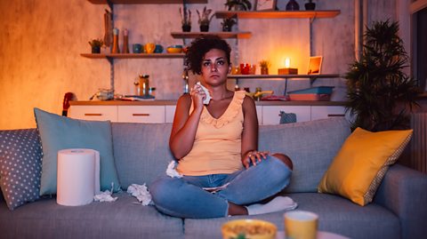 A person wipes tears with tissue whilst sat on their sofa watching a movie