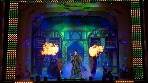 Colourful stage performance of Dick Whittington with villain dressed in dark green take centre stage featuring elaborate set design with Tudor-style houses, dramatic lighting, and fire effects