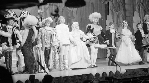 Vintage black-and-white photo of a theatrical cast in ornate costumes during a pantomime stage performance with period-style backdrop.