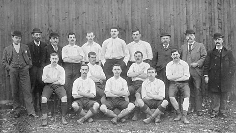 Historic black-and-white photograph of an early football team posing outdoors in front of a wooden fence, with players wearing vintage kits and knee-high socks, accompanied by men in formal suits and coats.
