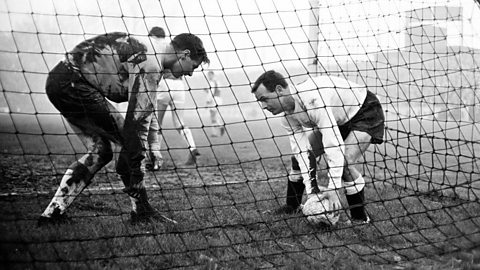 Historic black-and-white football photograph showing two players inside the goal area during a muddy match, one bending to pick up the ball while the other stands nearby, with the goal net in the foreground