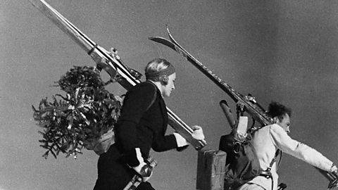 German couple climbing up a snow hill with skis, a christmas tree and parcels (1937)