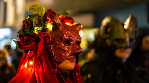 Close-up of a person wearing a red devil-style mask adorned with roses, greenery, and small glowing lights during a festive outdoor event, with another masked figure in dark costume visible in the background.