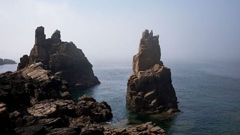 Rocky sea stacks rising from the clear blue water along a rugged coastline, with jagged formations and misty horizon creating a dramatic coastal landscape.