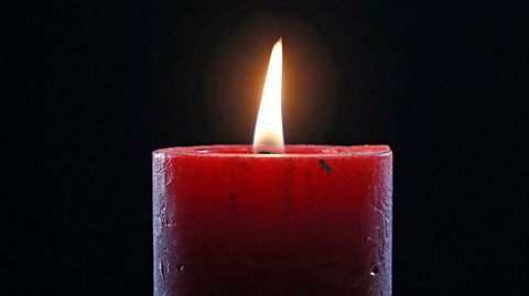 Close-up of a single red candle burning brightly against a dark background, surrounded by green pine branches, symbolizing a traditional Christmas decoration.