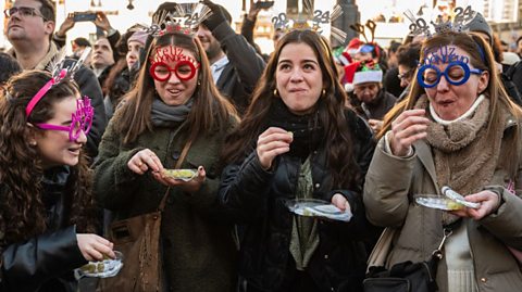 Four women in Spain eat a grape and make a wish for each chime of midnight on New Year's Eve, wearing 2024 party glasses