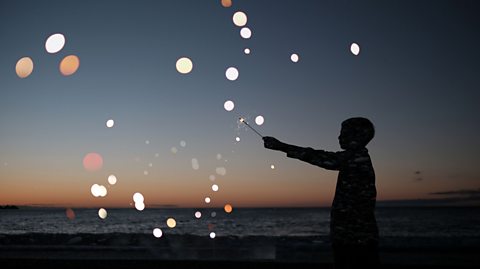 Silhouette of a person holding a sparkler on a beach at dusk, with glowing bokeh lights scattered across the scene and a calm ocean under a colorful twilight sky.