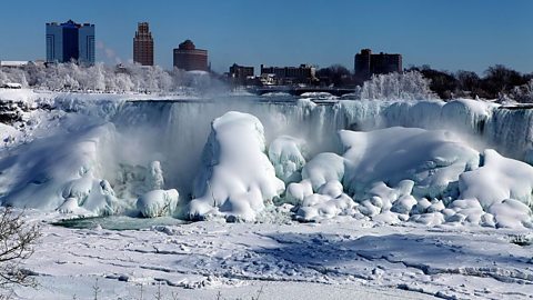 Large natural ice sculptures form at the base of cascading waterfall with a city skyline in the background