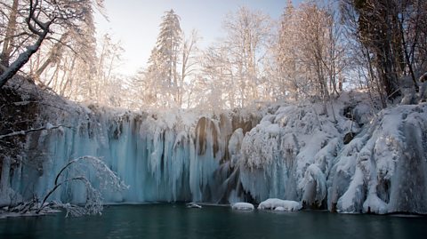 A lake in Plitvice Lakes National park glistens green in the sun, above it is a horsehoe shoaped frozen waterfall with the sun rising through the trees