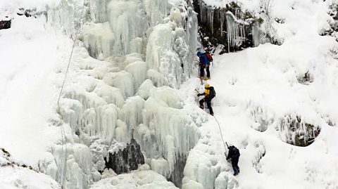 A group of ice-climbers scale up the side of a frozen waterfall in single file, avoiding icicles and other icy formations