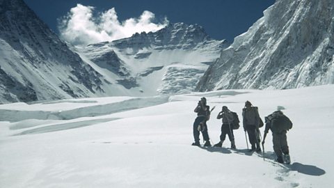 Mountaineers trekking across a snowy glacier toward towering Himalayan peaks under a clear blue sky. Climbers in winter gear with backpacks and ice axes ascend through deep snow in a remote high-altitude mountain landscape, surrounded by rugged cliffs and dramatic ice formations.