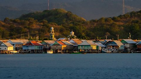 Colourful waterfront houses line the shore of a calm bay with lush green hills and tall communication towers in the background. Beyond the hills, a majestic mountain range rises under a dramatic sky with layered clouds, bathed in warm golden light from the setting sun. The scene captures a tranquil coastal village surrounded by natural beauty.