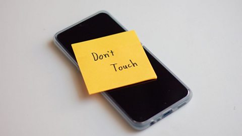 A smart phone is turned off with a bright orange note stuck on it saying 'don't touch'