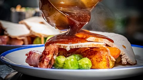 A ladle of gravy is poured onto a roast dinner on a white plate with a blue outline. On the roast is sprouts, potatoes, pigs in blankets and a slice of turkey
