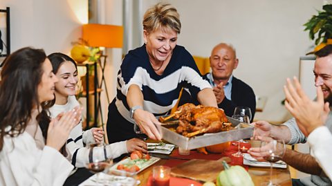 A family gathered around a dinner table with plates and glasses of wine applaud as a woman wearing a navy and white striped top puts a roast chicken in a silver tray in the middle of the table