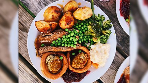 An aerial view of a roast dinner on a white plate. The dish includes two Yorkshire puddings, gravy, roast beef, peas, mashed potato, five roast potatoes and cranberry sauce