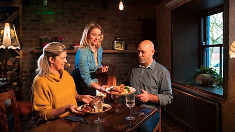 A couple sit a table inside a rustic pub, with vintage lamps and bare brick walls. A blonde woman wears a yellow jumper and a man wears a grey buttoned shirt as a lady in a denim top and black pants places roast dinners on their table
