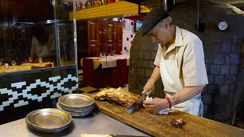 A man wearing a black flat cap and a cream shirt with an apron on chops mean on a wooden slab in a kitchen. He is making asado, a traditional Argentinian barbecue meal that heavily features beef as well as sides and vegetables