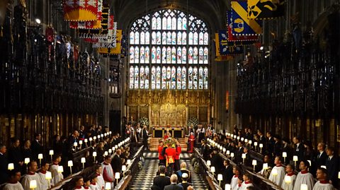 Familja Mbretërore ndjek arkivolin e Mbretëreshës Elizabeth II teksa transportohet në Kapelën e Shën Gjergjit, 2022. Kapela është plot me arkitekturë gotike, dritare me xham të pikturuar dhe flamuj. Djemtë e korit veshin të bardha në të dyja anët e korridorit dhe të afërmit dhe mysafirët veshin të zeza pranë altarit.