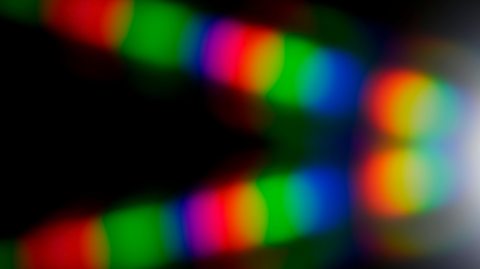 A vibrant abstract image featuring overlapping bands of bright colours of green, red, blue, and yellow—against a black background. The colors appear as soft, glowing streaks or blurred light patterns, creating a sense of motion and depth. On the right side, the light intensifies into a bright white glow, adding contrast to the colourful sections.