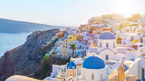 Santorini at sunset. The Aegean Sea surrounds the island and the sun sets on the rocks and the colourful houses on the cliffside, including white buildings with blue domes