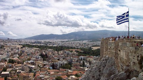 An aerial view of Athens, the capital of Greece. Greece's blue and white striped flag stands tall on the cliffside as tourists look out over the city at the buildings, ruins and greenery