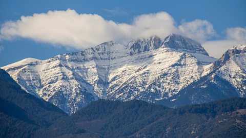 A view of Mount Olympus, Greece. The mountain is covered in a dusting of snow. Below is trees and above the mountain is clouds on a clear blue sky