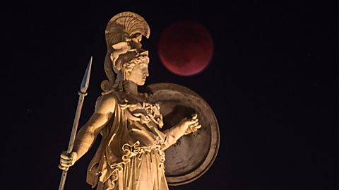 A statue of Athena, Greek goddess of wisdom and warfare, in Athens. The photo has been taken at night and behind the statue is the dark sky and a full moon, known as a blood moon, for its red hue during a lunar eclipse. The statue is marble and Athena wears a tall helmet, holds a spear and a shield and wears a long dress
