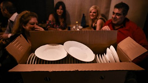 A cardboard box is full of white plates in a Greek restaurant ready to be smashed for a celebration. Behind the box is a woman and her partner laughing on one table, while the other table behind them has two women sitting down looking on curiously