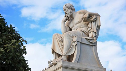 A statue of Greek philosopher Socrates in Athens. The statue is white marble and Socrates sits on a chair, wearing robes around him and sandals. He has a curly beard and curly hair and his chin rests on his right arm as he thinks