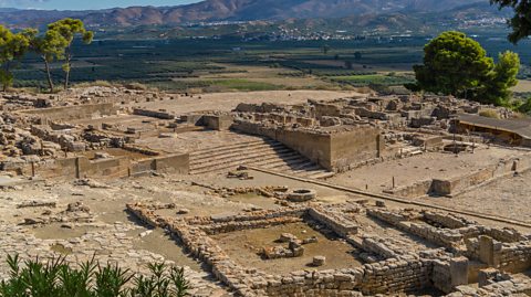 Ruins of the Phaistos Minoan Palace and village in Crete. The area is full of old slabs and bricks that still outline the buildings that once stood there. In the distance is greenery and houses