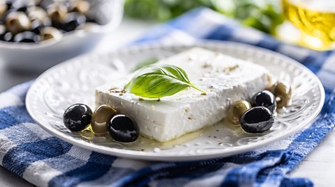 A large, rectangular cube of feta cheese, drizzled with olive oil and olives surrounding it. It is served on a white plate which is on top of a blue and white tea towel