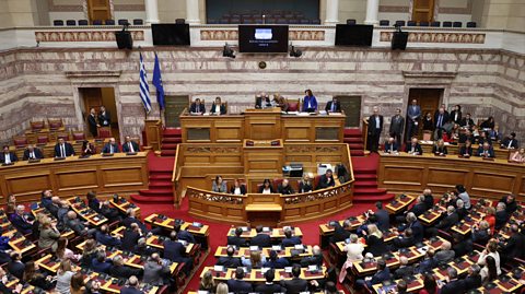 A view of the parliamentary session for the election of Konstantinos Tasoulas as Greece's new president, 2025. The aerial shot shows members of parliament on a semi-circle of benches in front of wooden stands where more members sit. The carpet is red and the grand hall has intricate stonework