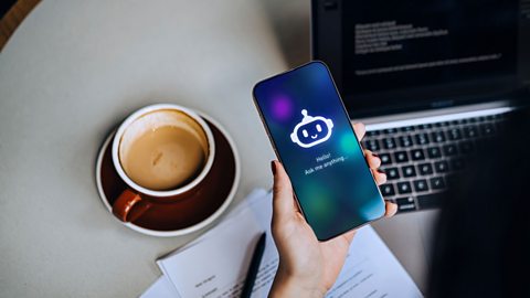 A hand holding a phone with a generic AI chatbot on it, with a laptop and coffee and paper notes in the background