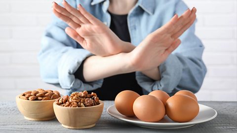 A woman wearing a black top and denim shirt crosses her hands. In front of her is a white plate with five eggs on it, as well as two wooden bowls next to it containing different nuts