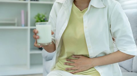 A woman sits wearing a light green top, white unbuttoned shirt and white pants. She holds her stomach with her left hand and in her right hand, she holds a glass of milk