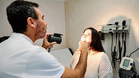 A GP wearing a white top sees to a patient, a woman with brown hair wearing a cream jumper sits on a bed in the office. She opens her mouth wide as the GP shines a light down her throat to check it