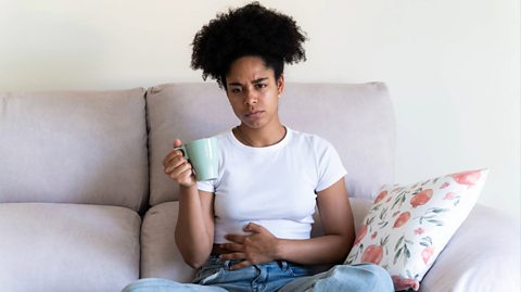 A woman with curly hair tied up sits on her grey couch wearing a white top and jeans. She looks uncomfortable as she clenches her stomach with her left arm and holds a light green mug in her right hand