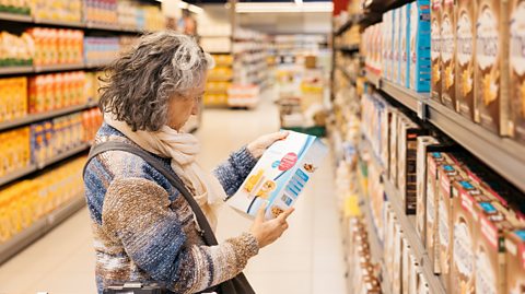 A woman with grey curly hair wearing a cream scarf and blue and neutral jumper stands in a supermarket aisle full of boxes of cereal. She holds one in her hand reading the box