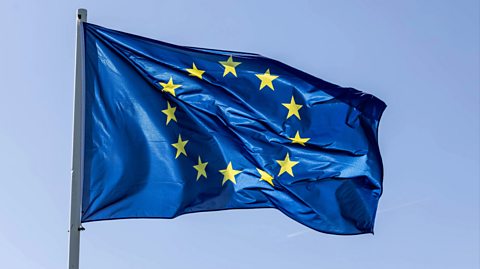 The European Union flag waves from a large silver pole. The flag is dark blue and has a circle of 12 yellow stars in the centre