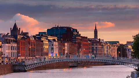 The Ha'Penny Bridge over the River Liffey in Dublin, Ireland. People walk over the bridge at sunset, with colourful buildings lined along the lefthand side of the river