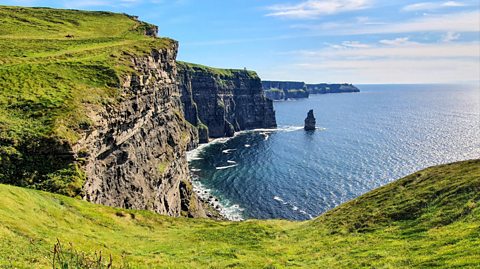 The green Cliffs of Moher look out onto the Atlantic coast of Ireland on a sunny day, with blue skies and few clouds