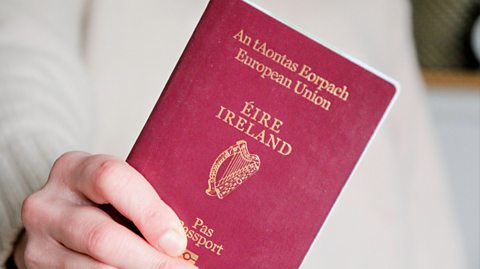 A hand holds an Irish passport, which is dark red and has gold writing, the main text reading 'Eire Ireland' with an illustration of a harp