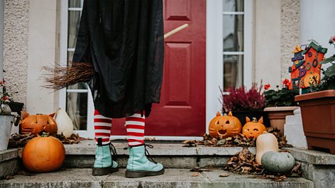 A young girl wearing light blue laced up boots, pink and red stripy tights, a black cape and a brooms stands on the front step of a house with a red door. On the door step is a number of pumpkins, some carved like cats with ears and whiskers