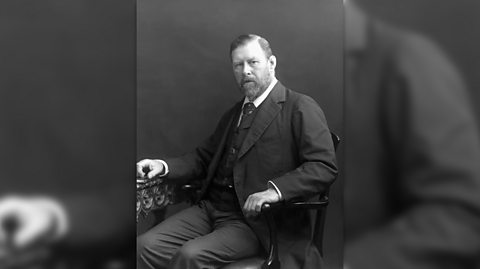 Author Bram Stoker famously wrote Dracula. Black and white image of Bram sitting on a wooden chair looking at the camera. He has a beard and short hair and wears and dark three-piece suit