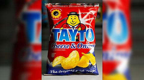 A packet of cheese and onion Tayto crisps, which are red with blue text and an illustration of a potato man wearing a red jacket and black hat and tie