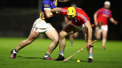 Two men, one wearing white shorts and a blue jersey and the other wearing white shorts, a red jersey and a helmet during a game of hurling. They are on a green pitch with a stick and ball