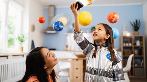 A mum with dark hair and a light orange top smiles and looks at her young daughter, who also has dark hair and is wearing a metallic silver space suit. The little girl is shining a light above her as model figures of the sun and Solar System planets hang from the ceiling 
