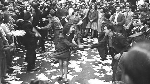 Crowd celebrating in a city street after World War 2, with people dancing among scattered paper and onlookers gathered around in festive atmosphere.